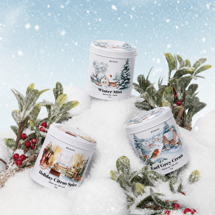 Three white cylindrical candles with illustrated winter labels are arranged in snow with frosted greenery and red berries. The labels read Pine Ridge and show scents Winter Mint, Holiday Chronicle, and East Coyote Cream.