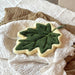 leaf shaped hand towel pictured on a stack of fabric. cream coloured edge with a dark green leaf.