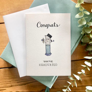 Graduation card with a cartoon graduate on a wooden surface with greenery