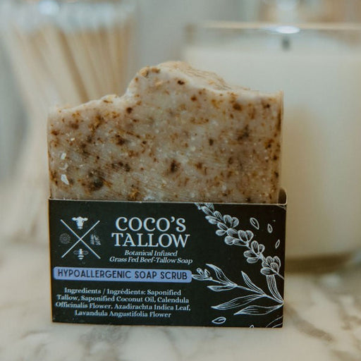 A bar of Coco's Tallow Hypoallergenic Soap Scrub in its black packaging box, set on a marble surface with a white candle and gold accessories blurred in the background.