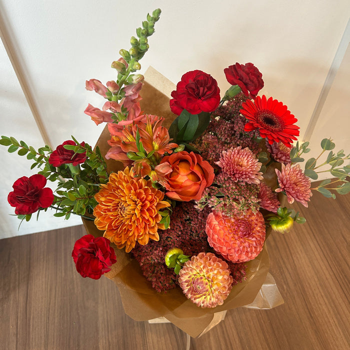 A vibrant autumn flower bouquet wrapped in brown kraft paper, featuring red carnations, orange dahlias, burgundy chrysanthemums, and green foliage, set on a wooden floor against a plain wall.