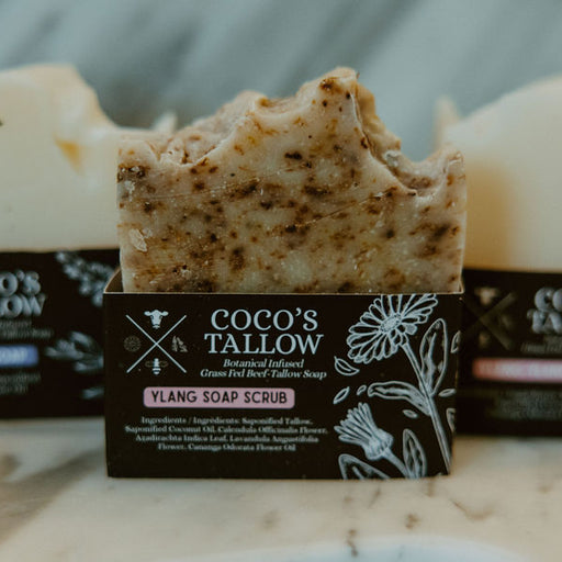 Three bars of Coco's Tallow Ylang Soap Scrub arranged on a marble surface with their dark brown paper packaging in the background.