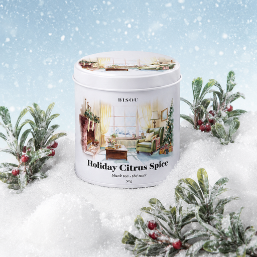 A white cylindrical tin of BISOU Holiday Citrus Spice sits in a snowy winter scene with frosted greenery and red berries. The tin features a festive illustration of a cozy living room with a Christmas tree.