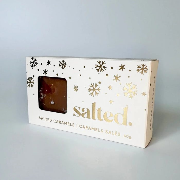 A rectangular box of 'salted.' brand salted caramels with a clear window showing the brown caramel product inside, decorated with gold snowflakes and text on a light gray background.