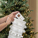 A white knit Christmas stocking with fringe, pom-poms, and a striped pattern hangs from a green Christmas tree decorated with warm yellow lights. A person's hands are shown holding a packaged gift inside the stocking.
