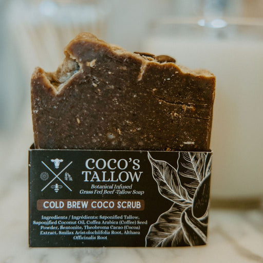 A bar of Coco's Tallow Cold Brew Coco Scrub soap standing upright in its branded cardboard packaging, photographed on a light marble-like surface with a softly blurred bathroom or kitchen background.