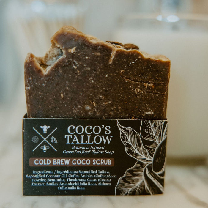A bar of Coco's Tallow Cold Brew Coco Scrub soap standing upright in its branded cardboard packaging, photographed on a light marble-like surface with a softly blurred bathroom or kitchen background.