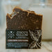 A bar of Coco's Tallow Cold Brew Coco Scrub soap standing upright in its branded cardboard packaging, photographed on a light marble-like surface with a softly blurred bathroom or kitchen background.