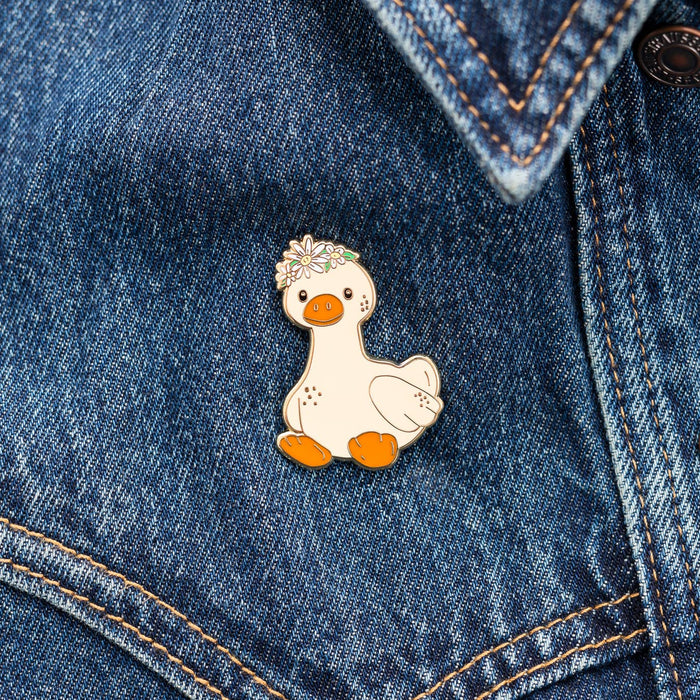 A small white duck enamel pin with orange feet and beak, wearing a floral crown, pinned onto a blue denim jacket.