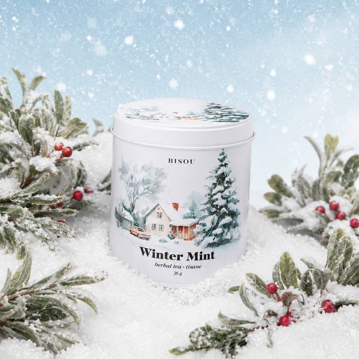 A white cylindrical tin of BISOT Winter Mint herbal tea sits in a snowy, festive setting with snow-covered pine branches and red berries against a light blue winter sky background.