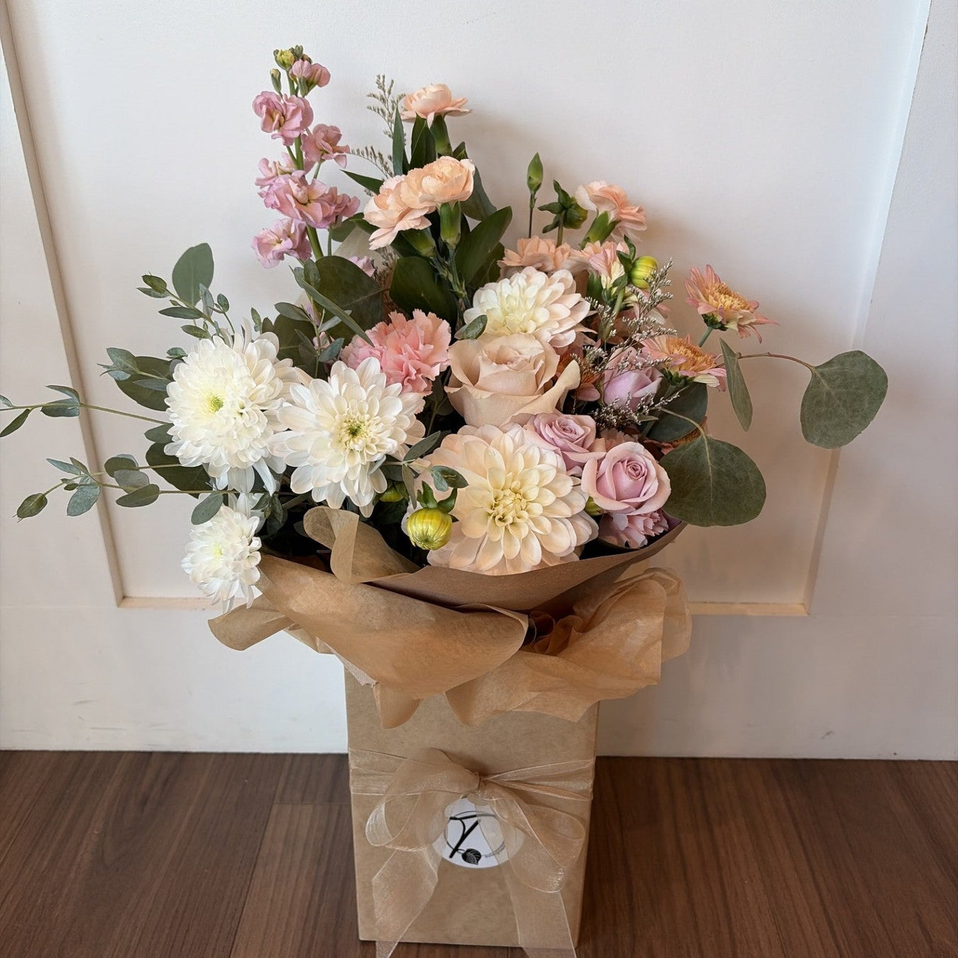 A floral arrangement in a kraft paper bag on a wooden floor against a white paneled wall. The bouquet contains white and pink flowers with green foliage, wrapped in brown paper with a small round logo tag.
