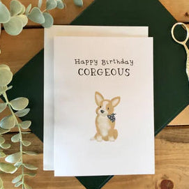 Birthday card with a corgi dog illustration and 'Happy Birthday CORGEOUS' text on a wooden surface with eucalyptus leaves and scissors.
