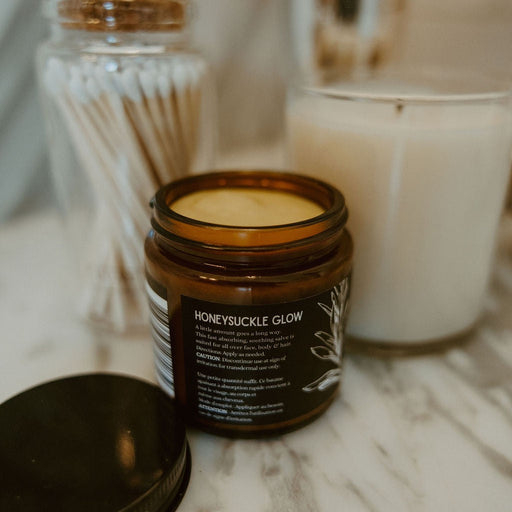 An amber glass jar candle labeled 'HONEYSUCKLE GLOW' with its black lid placed in front, styled on a marble surface with a jar of cotton swabs and a white candle in the background.