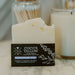 A bar of Coco's Tallow natural unrefined soap with a black label stands in front of a white candle on a marble surface, with cotton wipes and a gold bowl blurred in the background.