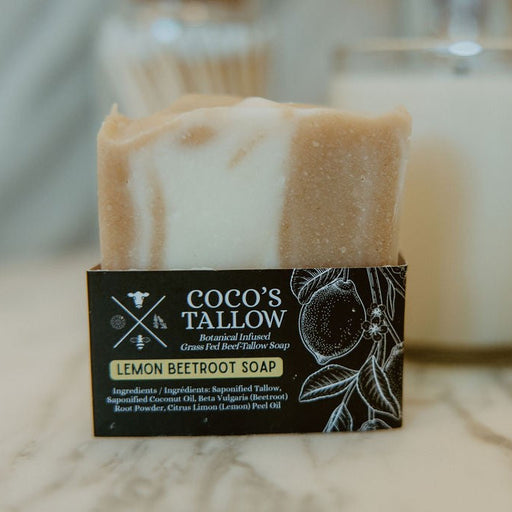 A bar of Coco's Tallow Lemon Beetroot Soap standing upright in its black cardboard packaging, with a blurred white candle in the background on a light marble surface.