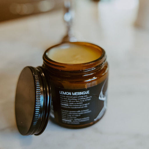 An open amber glass jar candle with its lid leaning beside it on a light marble surface. The label reads 'LEMON MERINGUE' and the candle wax is visible inside.