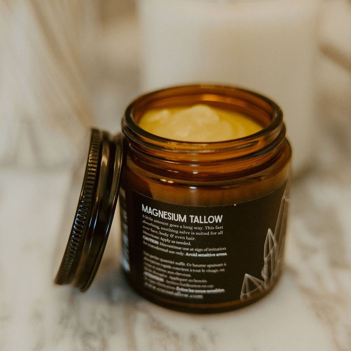 An open amber glass jar of Magnesium Tallow body butter sits on a marble surface with its lid leaning beside it. A white candle and other soft-focus household items are blurred in the background.