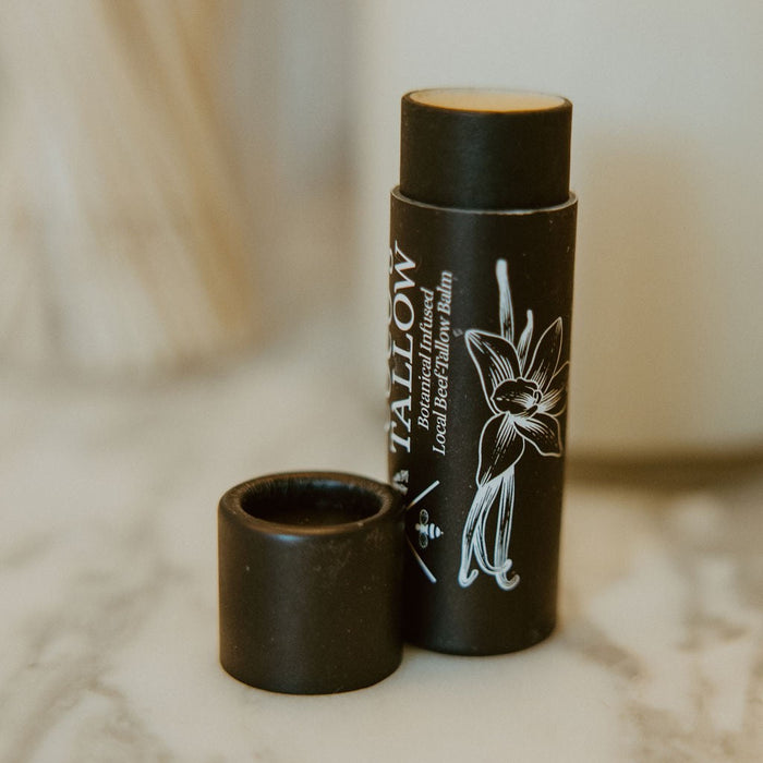 A dark brown lip balm stick labeled 'SALLOW' with its cap removed, standing on a light marble surface with a softly blurred background.