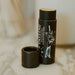 A dark brown lip balm stick labeled 'SALLOW' with its cap removed, standing on a light marble surface with a softly blurred background.