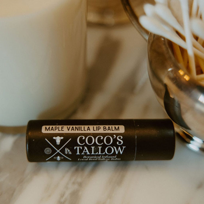 A black tube of Coco's Tallow Maple Vanilla Lip Balm resting on a white marble surface, styled with a white candle and cotton swabs in the background.