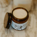 An open amber glass jar candle with cream-colored wax sits on a light marble surface, with a larger blurred candle in the background.