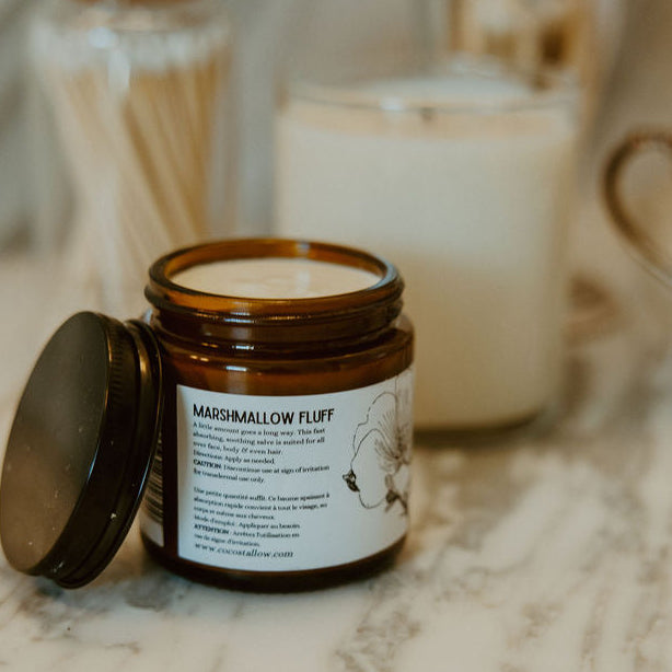 An amber glass jar candle with its lid off, labeled 'Marshmallow Fluff' by The Art of Scents, set on a marble surface with other candles in the background.