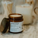 An amber glass jar candle with its lid off, labeled 'Marshmallow Fluff' by The Art of Scents, set on a marble surface with other candles in the background.