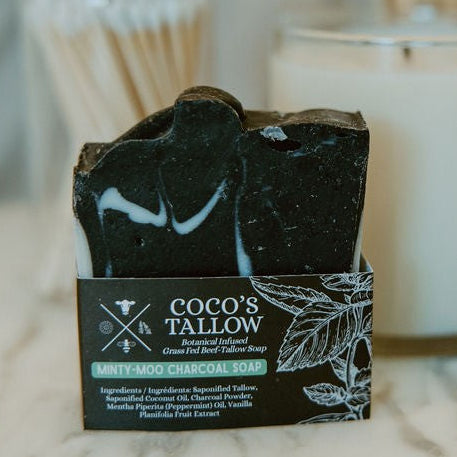 A bar of black marbled soap labeled 'COCO'S & TALLOW' stands on a marble surface with a candle, jars, and a gold cup blurred in the background.