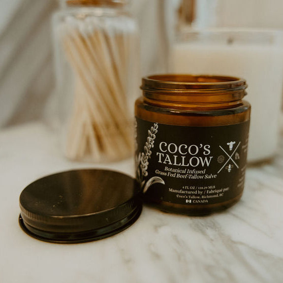 An amber glass jar of Coco's Tallow Beeswax Candles with its lid placed beside it on a white marble surface, with a softly blurred bathroom or vanity background containing a candle, cotton swabs, and a gold-toned object.