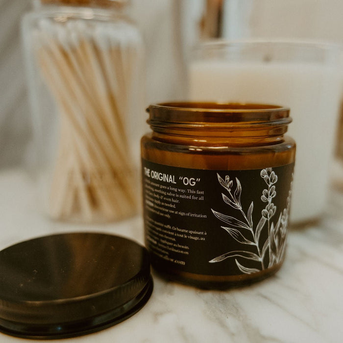 An amber glass jar candle with a black label and white botanical illustration, shown open with its lid placed beside it on a marble surface. A jar of cotton swabs and another candle are blurred in the background.