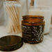 An amber glass jar candle with a black label and white botanical illustration, shown open with its lid placed beside it on a marble surface. A jar of cotton swabs and another candle are blurred in the background.