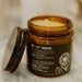 An amber glass jar candle with its lid off, showing a pale yellow wax inside. The jar has a dark brown label with white text that reads 'THE "OG" WHIPPED' and botanical illustrations.