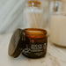 An amber glass jar of Coco's Tallow Shea Butter with its lid off, displayed on a marble surface with a blurred background showing a candle and dried flowers.