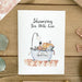 Greeting card with illustration of a bathtub and text 'Showering You with Love' on a wooden surface with decorative items.