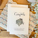 Graduation Greeting card with 'Congrats' and artichoke illustration on a wooden surface with decorative elements.