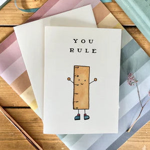 Greeting card with 'YOU RULE' text and cartoon character on a wooden surface with envelopes and a pen.
