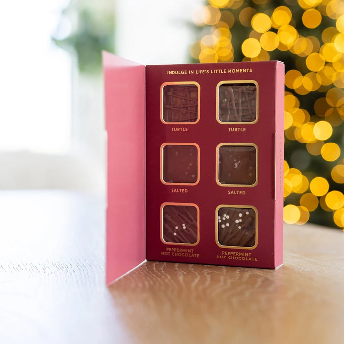 A pink and burgundy chocolate gift box labeled 'Indulge in Life's Little Moments' stands open on a wooden surface, with six square chocolate windows visible and a blurred Christmas tree with warm lights in the background.