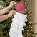 A white knit Christmas stocking with fringe and pom-pom details hangs in front of a lit Christmas tree. Two hands hold a pink greeting card inserted into the stocking.