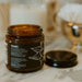 An open amber glass jar labeled 'TRAMSU' sits on a marble surface with its black lid beside it. The background is softly blurred, showing a white candle and other bathroom-like items in warm neutral tones.