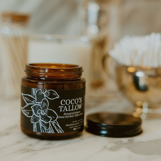 An amber glass jar candle labeled 'COCO'S TALLON' sits open on a light marble surface with its black lid beside it. The background is softly blurred with gold-toned decor and white cotton items.