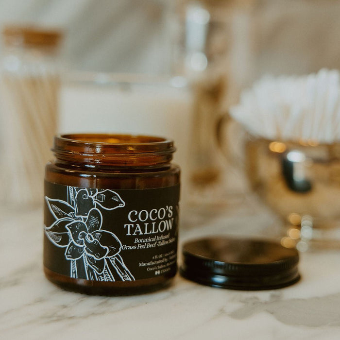 An amber glass jar candle labeled 'COCO'S TALLON' sits open on a light marble surface with its black lid beside it. The background is softly blurred with gold-toned decor and white cotton items.