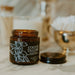 An amber glass jar candle labeled 'COCO'S TALLON' sits open on a light marble surface with its black lid beside it. The background is softly blurred with gold-toned decor and white cotton items.