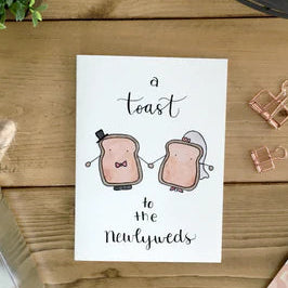 Newlywed Card with toast illustrations and text on a wooden surface with stationery items.