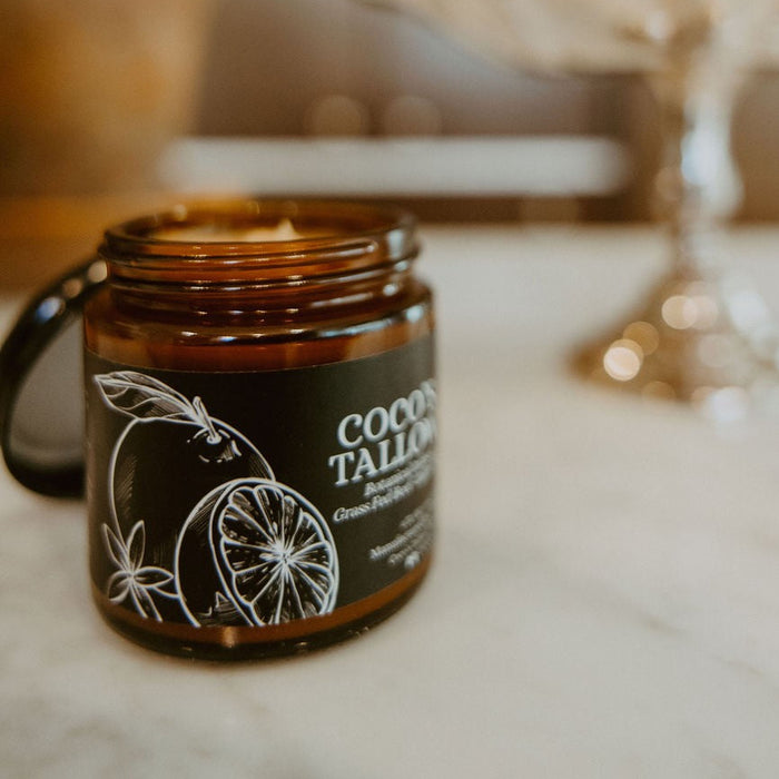 An amber glass jar candle with a black label reading 'COOCH TALON' and lemon illustrations, placed on a marble surface with lemons and a silver stand blurred in the background.
