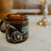 An amber glass jar candle with a black label reading 'COOCH TALON' and lemon illustrations, placed on a marble surface with lemons and a silver stand blurred in the background.