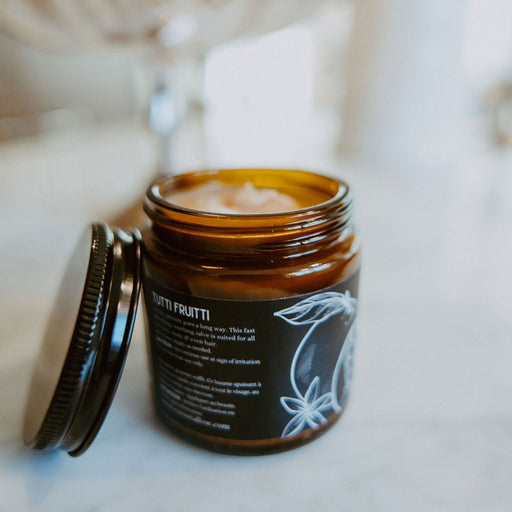 An amber glass jar of Nutri Fruitti body butter with its lid off, resting on a light marble surface with a softly blurred background.