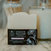 A white bar of soap in a black cardboard sleeve labeled 'COCO'S TALLOW' sits on a marble countertop, with a softly blurred white candle and bathroom accessories in the background.